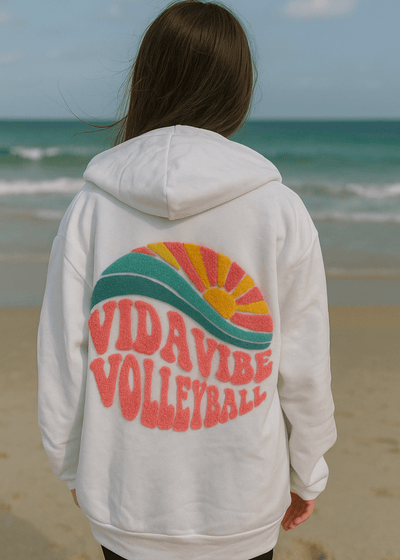 Person wearing a hoodie with 'Vida Vibes Volleyball' design on a beach